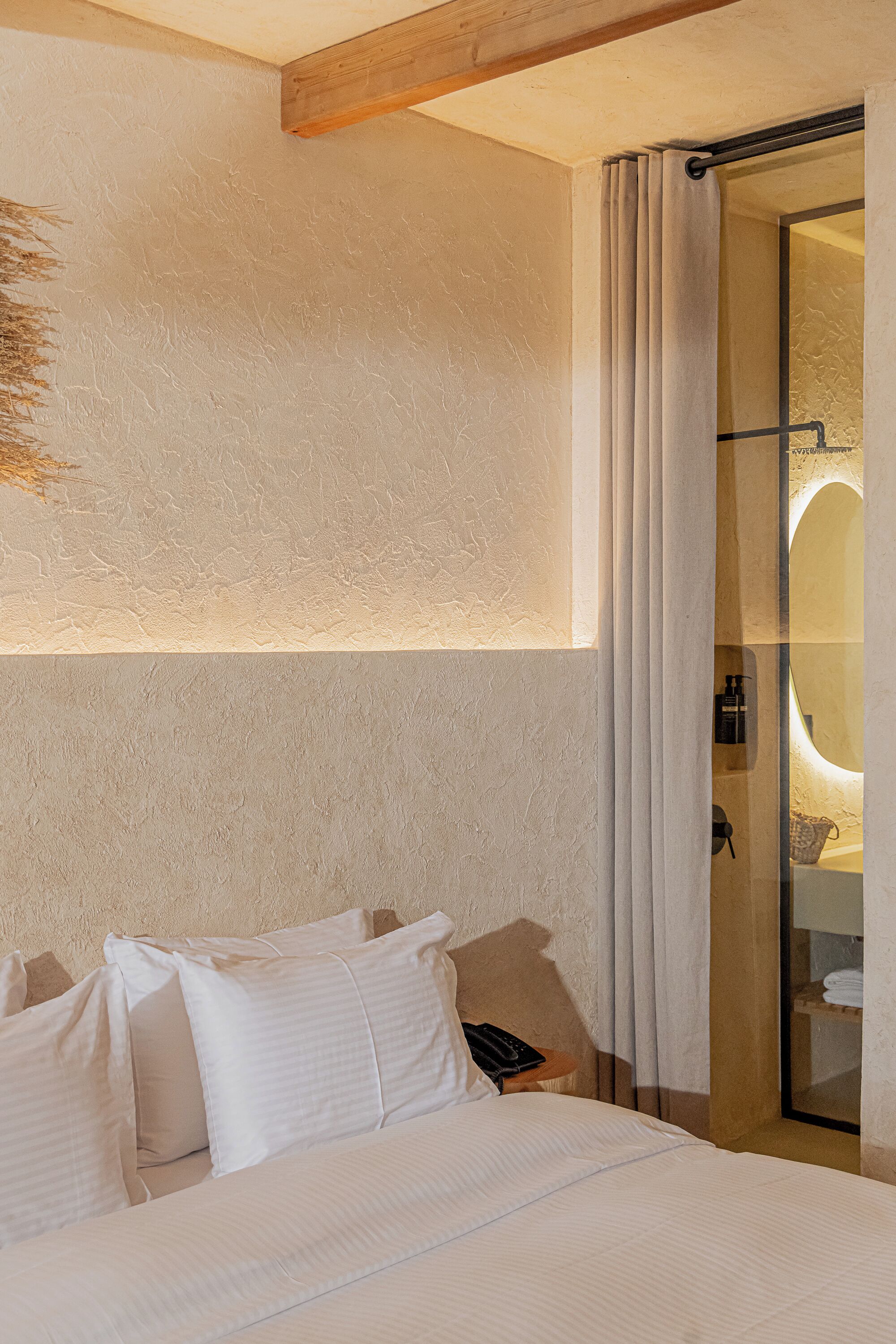 Un dormitorio con una cama grande, una viga de techo de madera, una cabina de ducha de vidrio y una lámpara fijada a la pared.