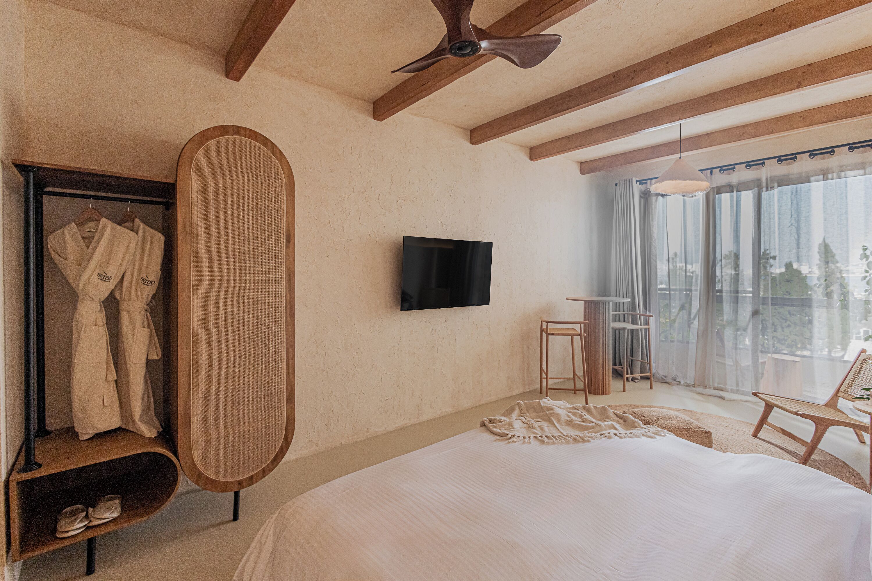 Un dormitorio con una cama grande, un televisor montado en la pared, un ventilador de techo, un armario con batas y un balcón con una silla.