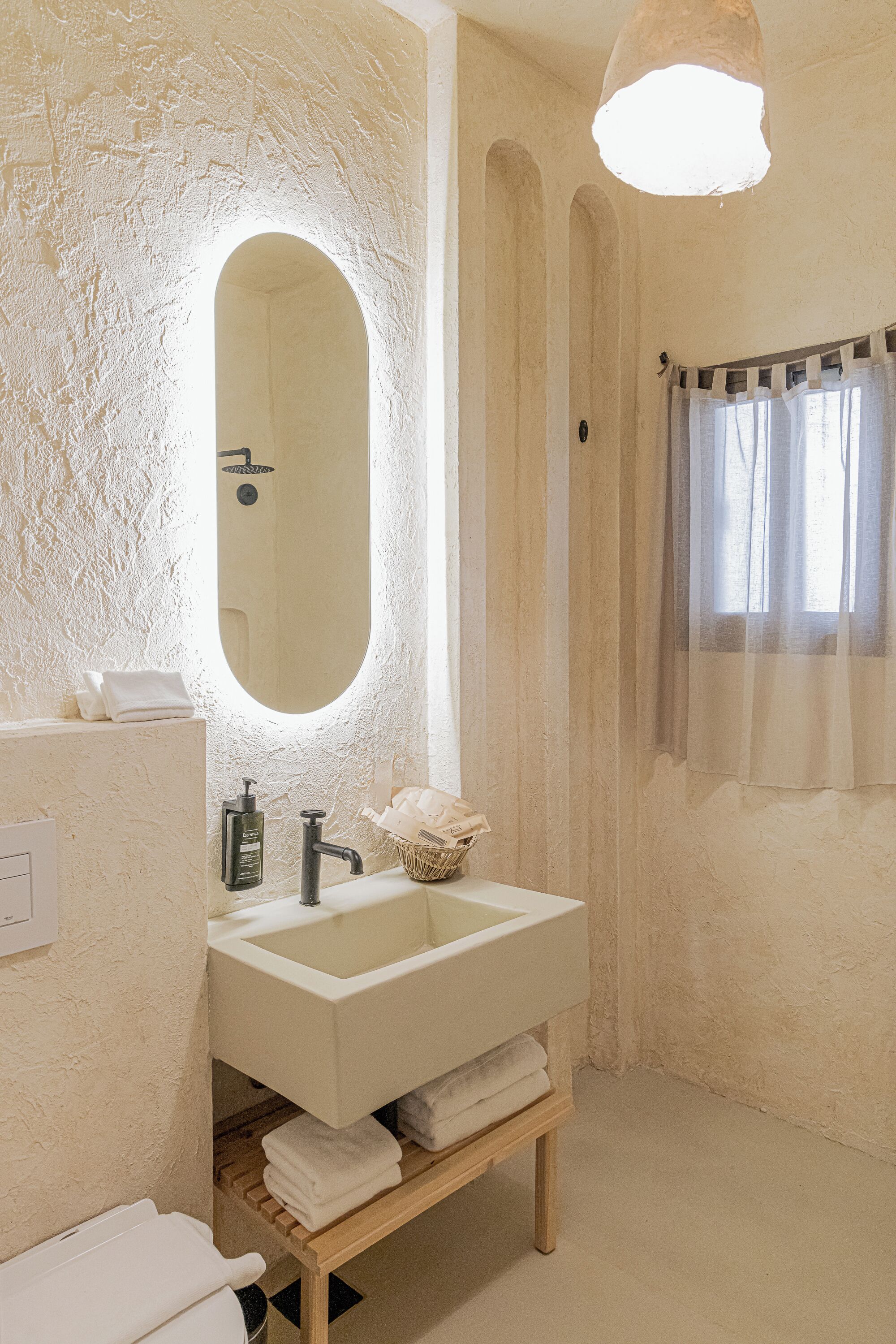 A bathroom with a large oval mirror, a white sink, a wooden shelf with towels, and a shower area with a curtain.