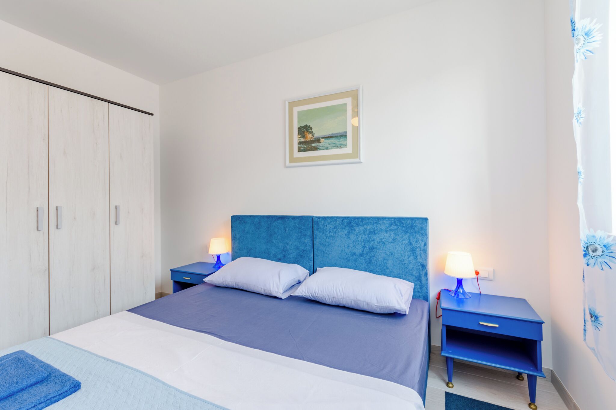 A bedroom with a bed, two bedside tables with lamps, a wardrobe, and a framed picture on the wall.