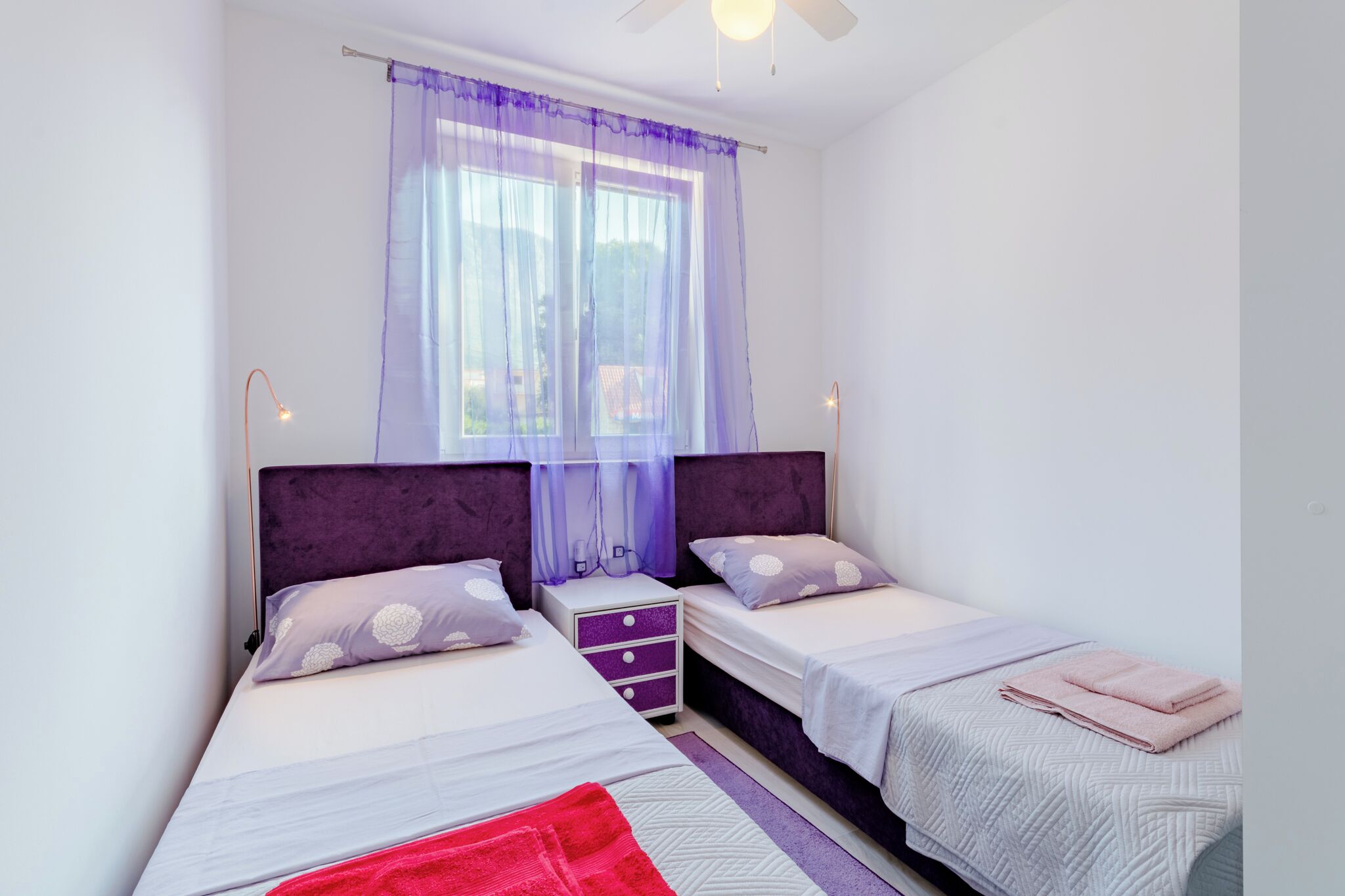 A bedroom with two beds, purple headboards, a window with sheer curtains, and a ceiling fan.