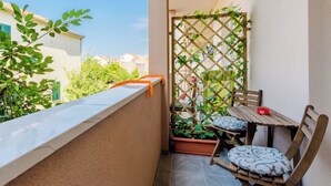 A small balcony with a wooden table and two chairs, a lattice screen with plants, and a view of neighboring buildings. - Apartment Karlo (Kastela)