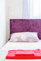 Una cama con cabecera morada, ropa de cama blanca y una manta roja.