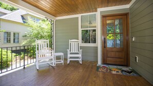 A porch with wooden flooring, a white rocking chair, a small table, and a wooden door.