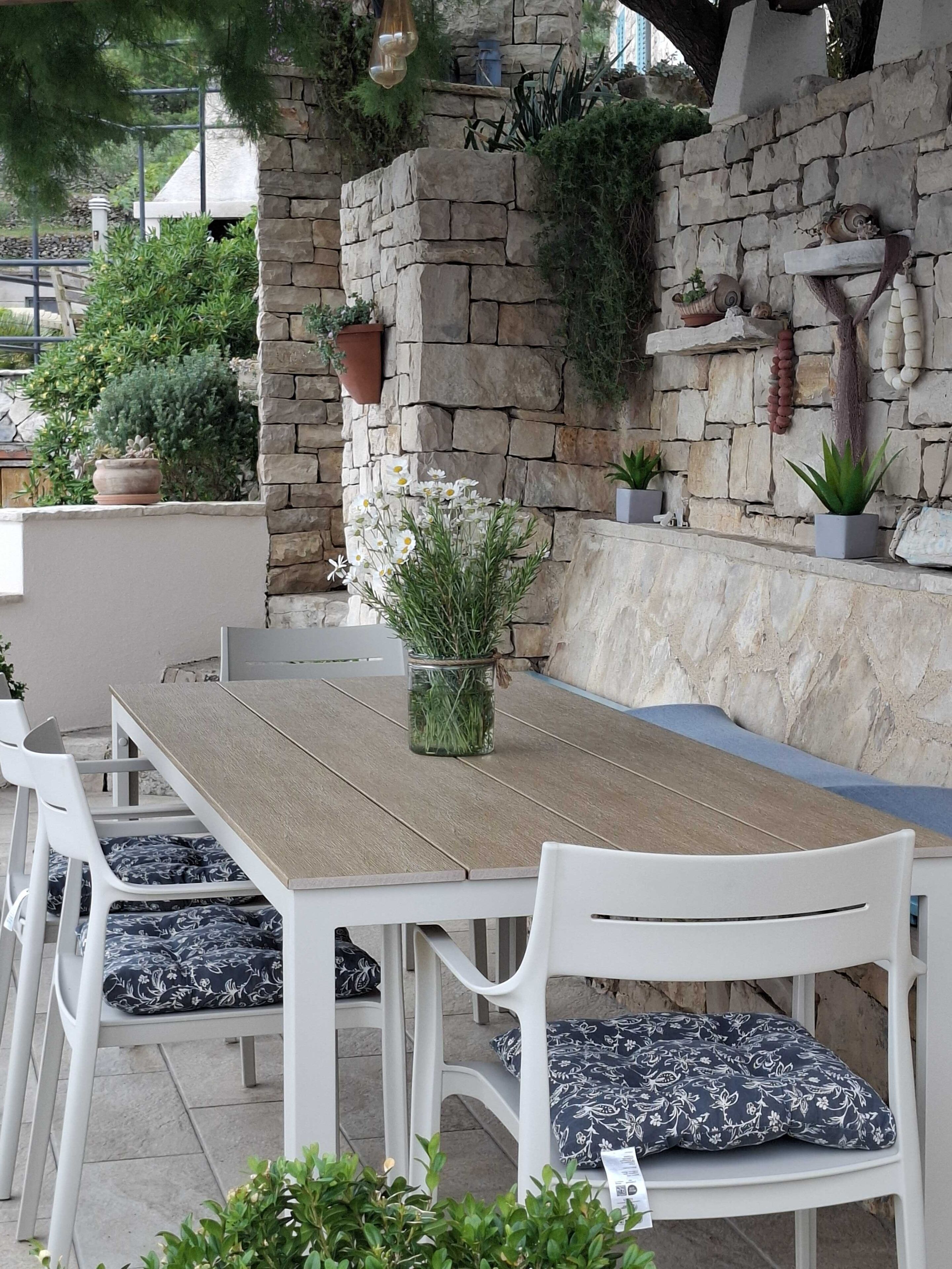 Un área de comedor al aire libre con pared de piedra, plantas en macetas y una mesa de madera.