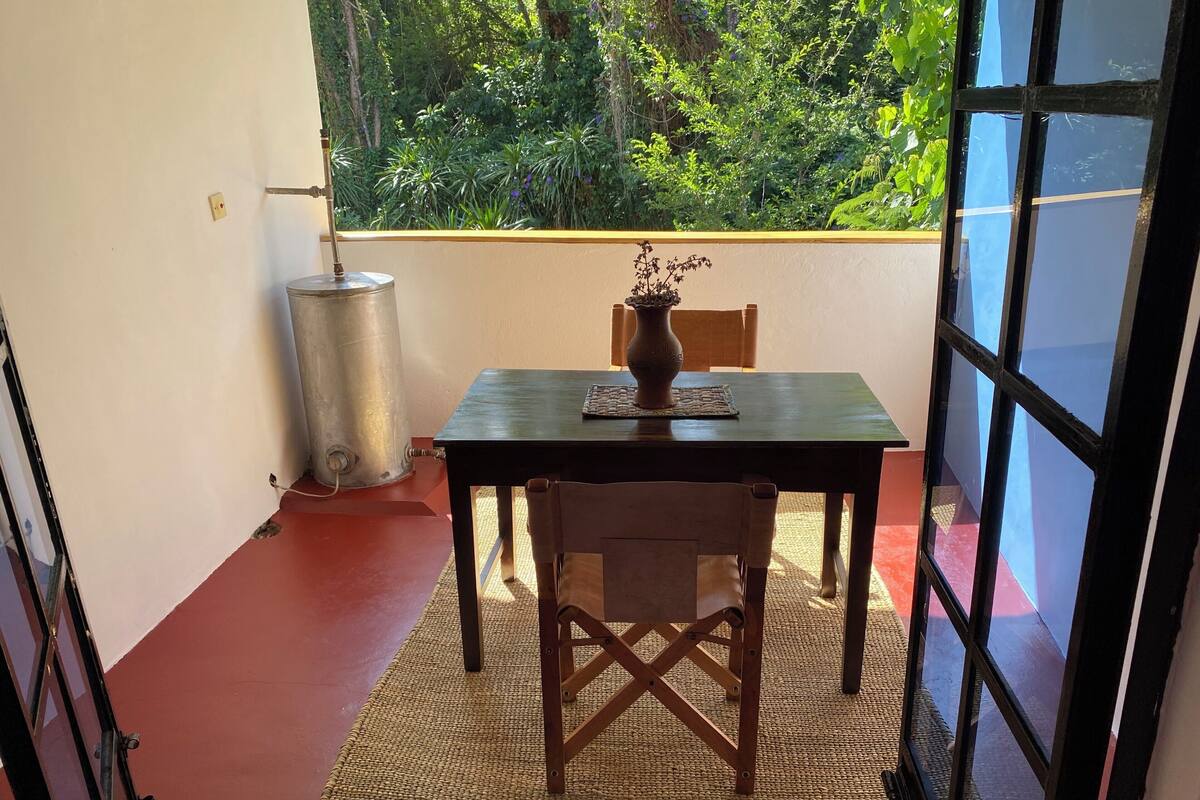 Una habitación pequeña con una mesa, una silla, un ventanal con vistas a la vegetación y un calentador de agua metálico.
