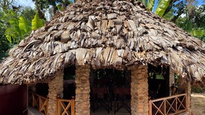 A thatched-roof hut with stone pillars and wooden railings, surrounded by a red floor and stone walls.