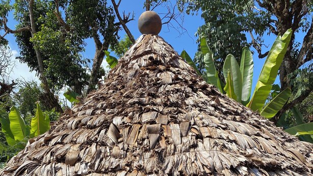 A thatched-roof structure with stone walls, a small balcony, and a red ground.