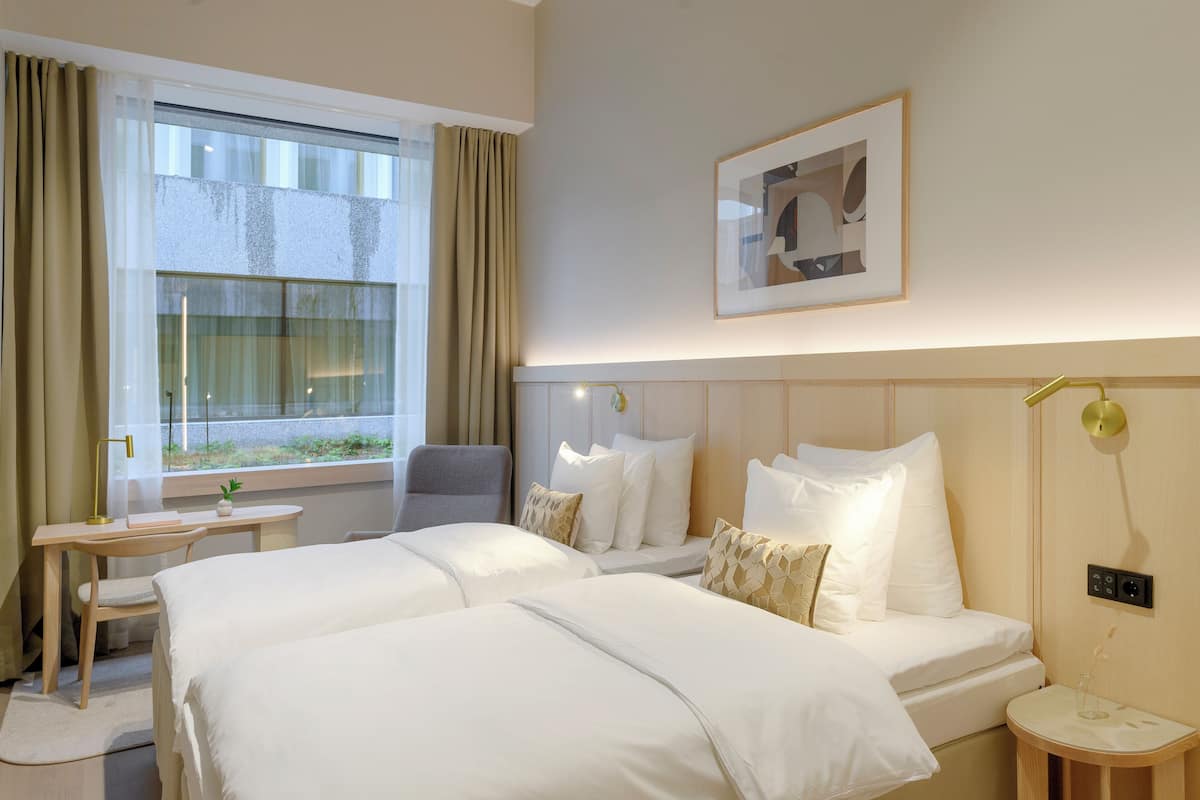 A hotel room with two beds, a desk, a chair, a window with curtains, a framed picture on the wall, and a bedside table with a lamp.