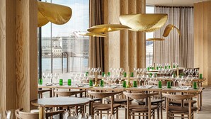Un restaurante con varias mesas dispuestas para comer, amplias ventanas y lámparas doradas colgantes.