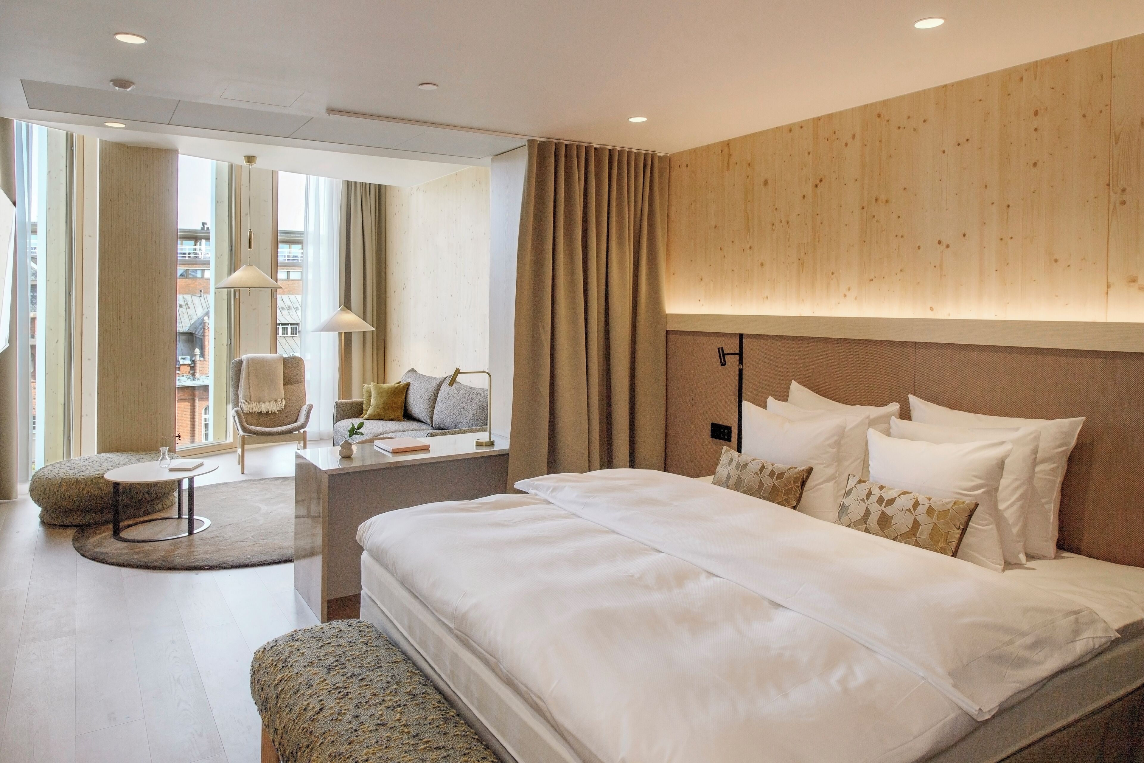 A modern hotel room with a large bed, a round coffee table, a chair, and a view of the city.