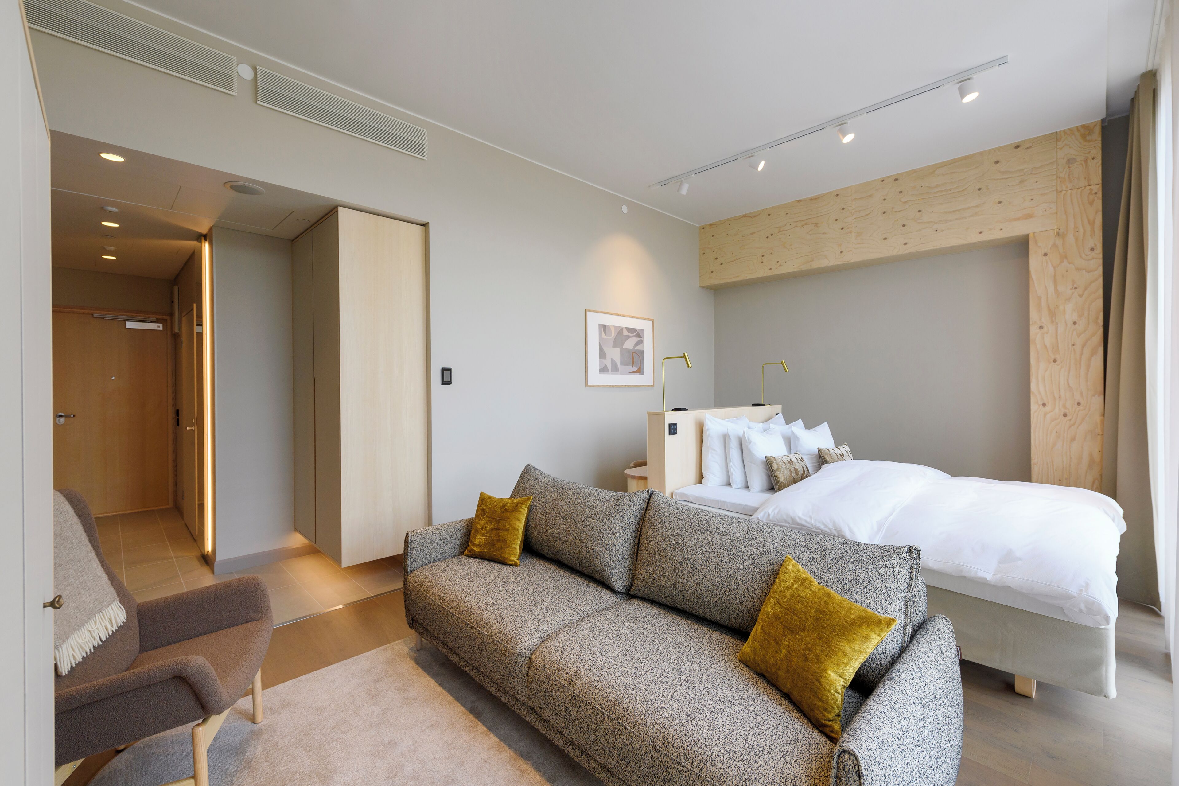 Une chambre d’hôtel moderne équipée d’un canapé, d’un lit, d’une table de chevet et d’une porte donnant sur un couloir.