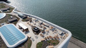 Vista aérea de una propiedad frente al mar con una terraza curva, zonas de estar al aire libre y una amplia piscina cubierta con vidrio.