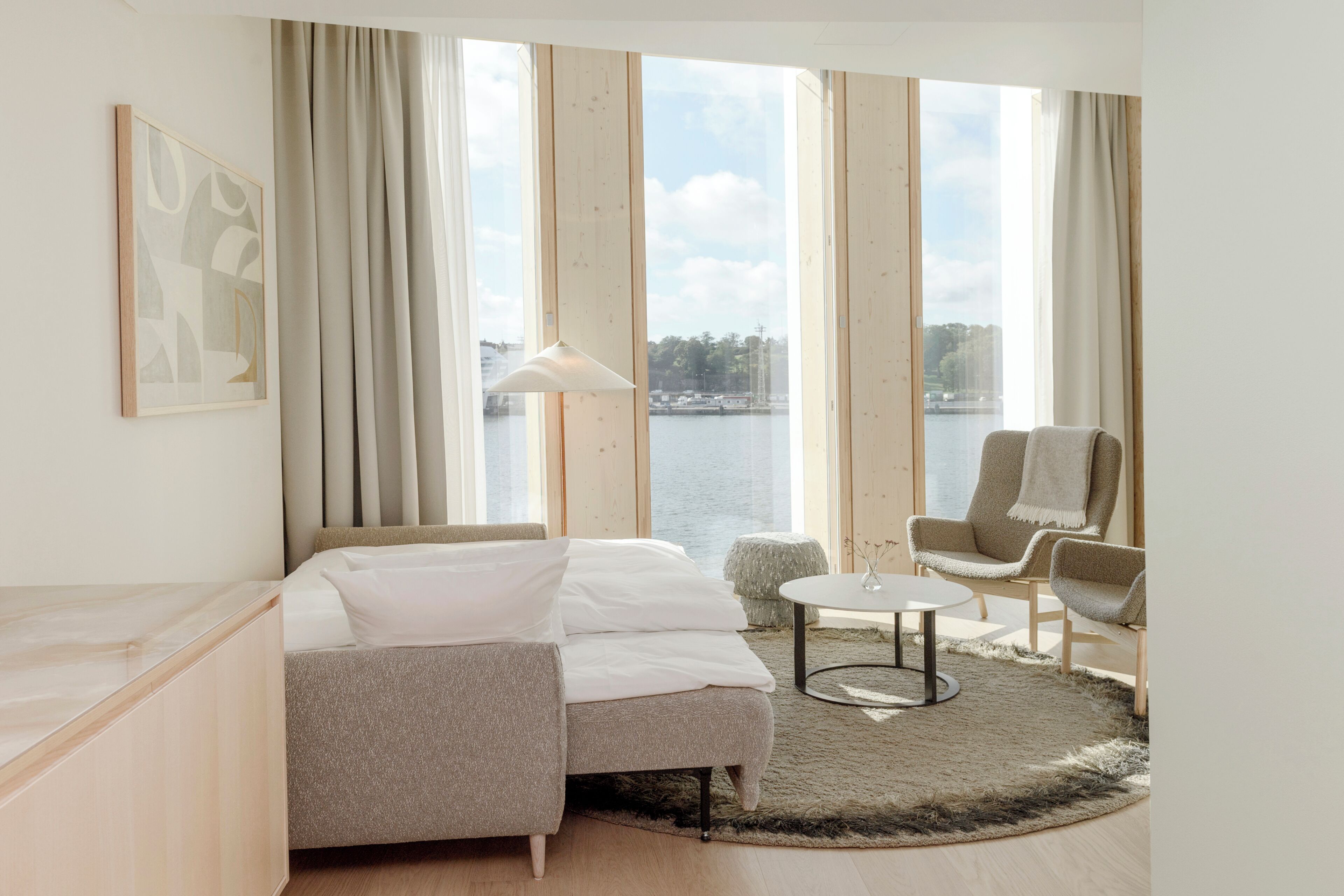 Hotelkamer met een bed, een stoel, een klein tafeltje en uitzicht op een waterpartij door grote ramen.