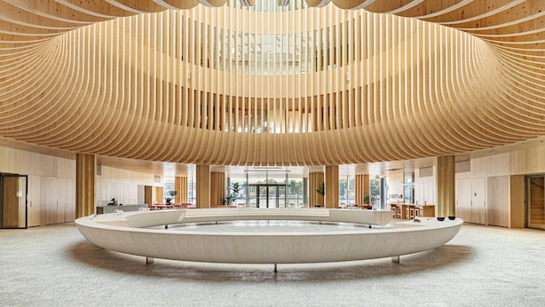 Un vestíbulo amplio con una zona de estar circular central, techo de listones de madera y grandes ventanales que aportan luz natural.