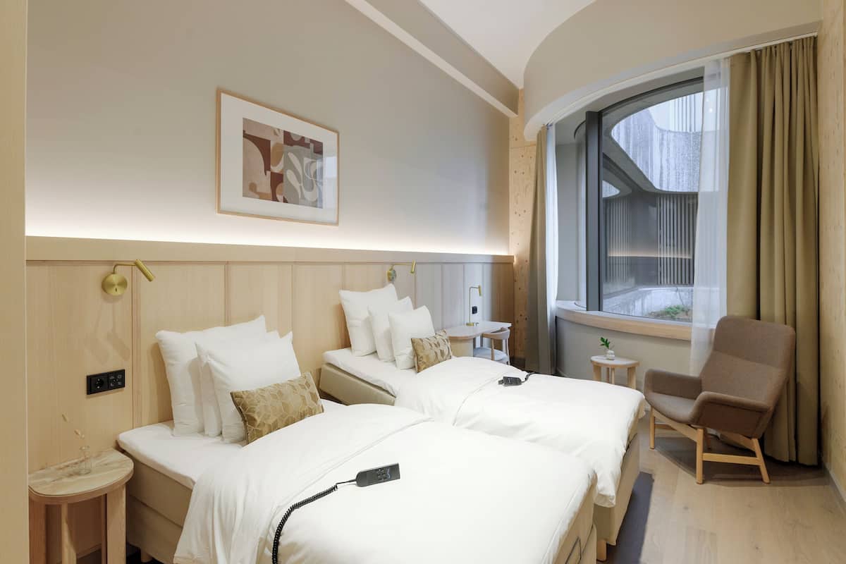 A hotel room with two beds, a chair, a small table, and a window with curtains.