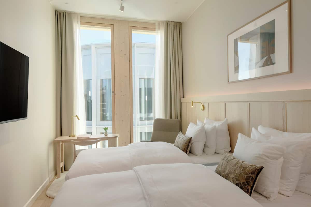 A hotel room with two beds, a desk, a chair, a TV, and a large window with curtains.