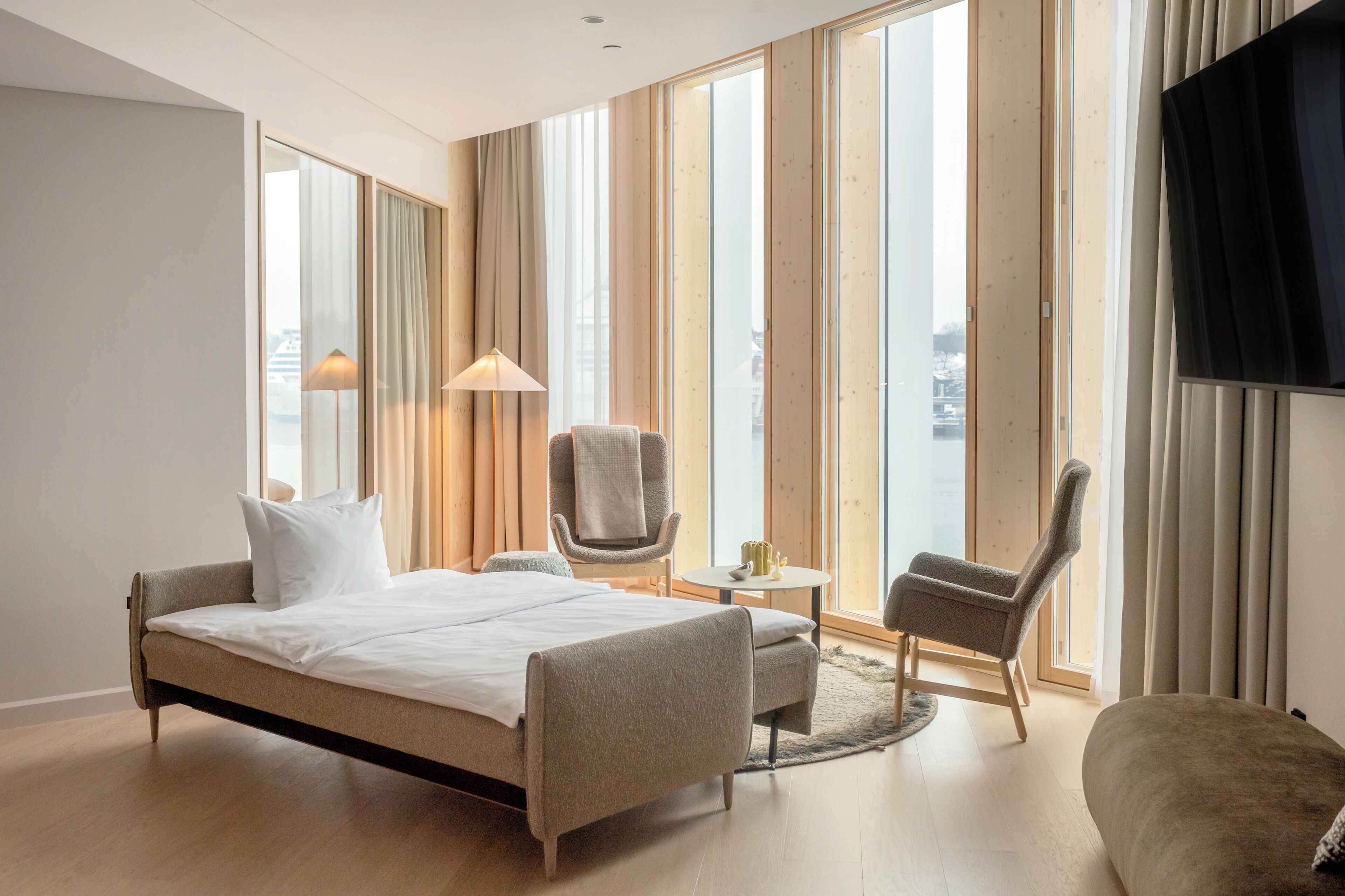 A modern hotel room with a bed, two armchairs, a small table, and a TV.