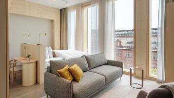 A modern hotel room with a sofa, a bed, a desk, and large windows with a city view.