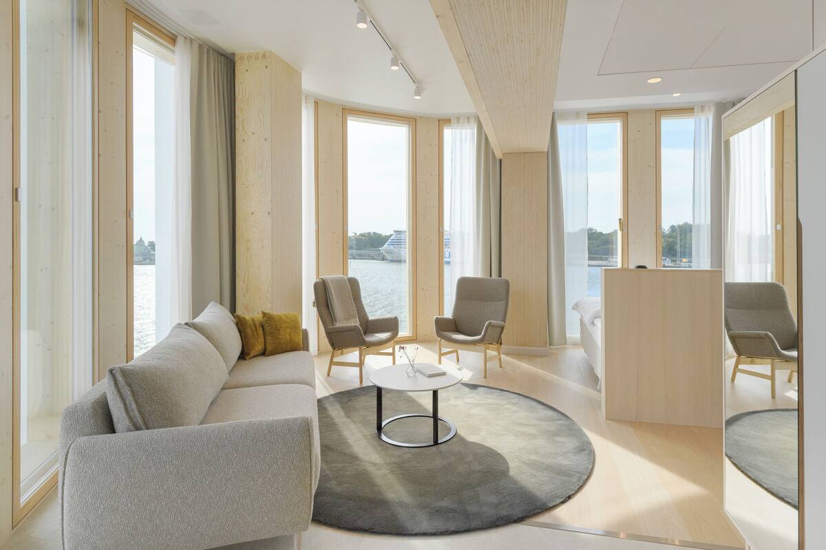 A modern living room with a sofa, chairs, a round coffee table, and large windows offering a view of a body of water.