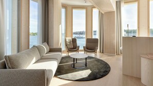 Una sala de estar moderna con un sofá, dos sillones, una mesa de centro y ventanas amplias que ofrecen vistas a un cuerpo de agua y al horizonte de la ciudad.