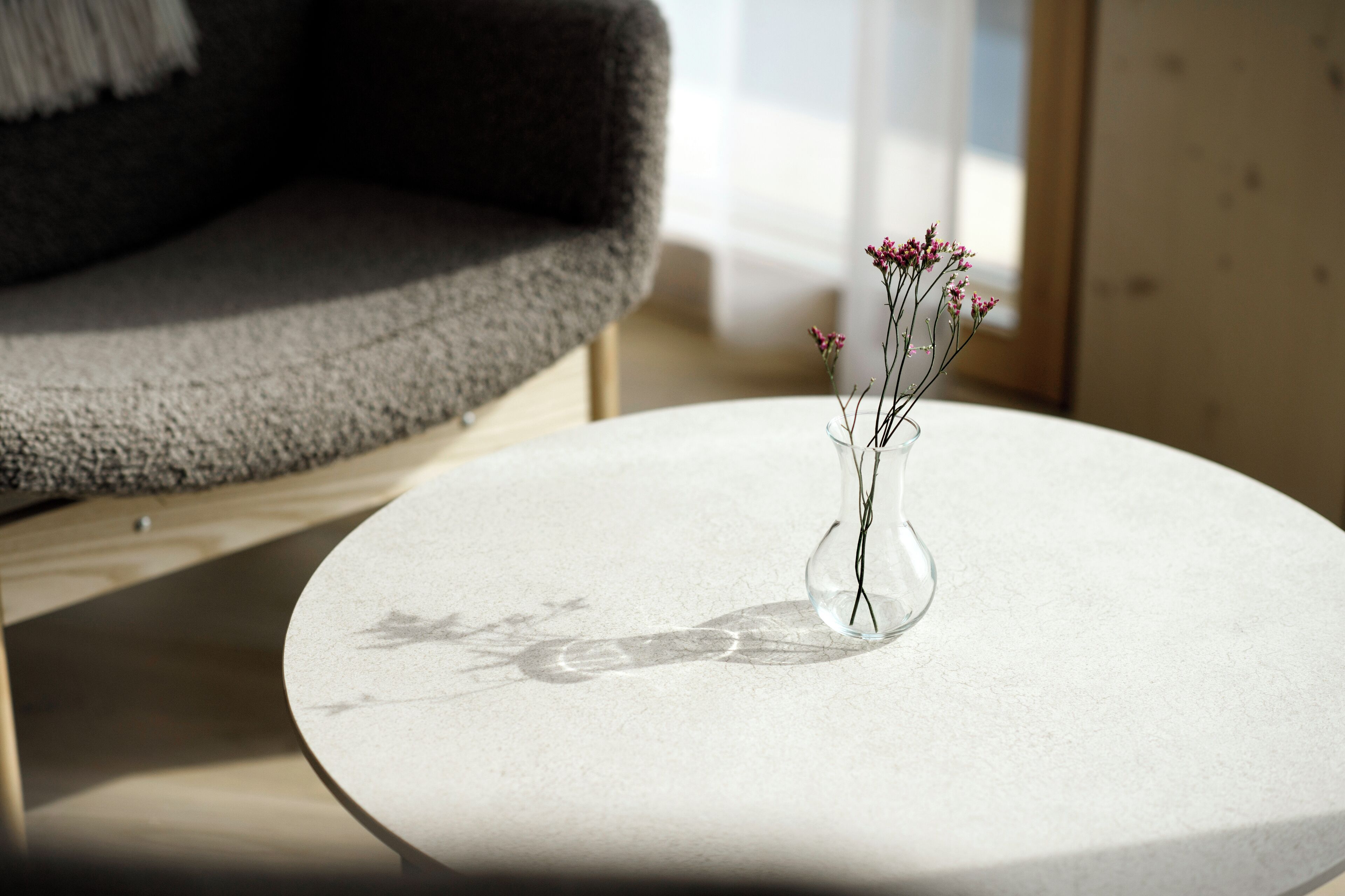 A round table with a clear vase containing flowers, positioned near a grey sofa and a window.