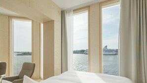 Habitación de hotel con una cama grande, dos sillones y vista a un crucero en el agua a través de amplias ventanas.
