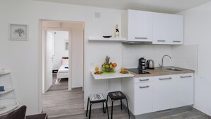 A modern kitchen with white cabinets, a wooden countertop, and a built-in stove. There is a fruit bowl with apples and oranges on the counter, and a bottle of champagne on a shelf above the sink. - Apartment Semi (Dubrovnik)