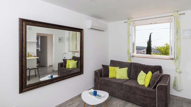 A modern living room with a large mirror, a brown sofa with yellow cushions, a small white table, and a window with a view. - Apartment Semi (Dubrovnik)