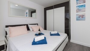 A neatly made bed with a white and blue striped bedspread, a pair of folded towels, and a bottle of wine on a tray. - Apartment Semi (Dubrovnik)