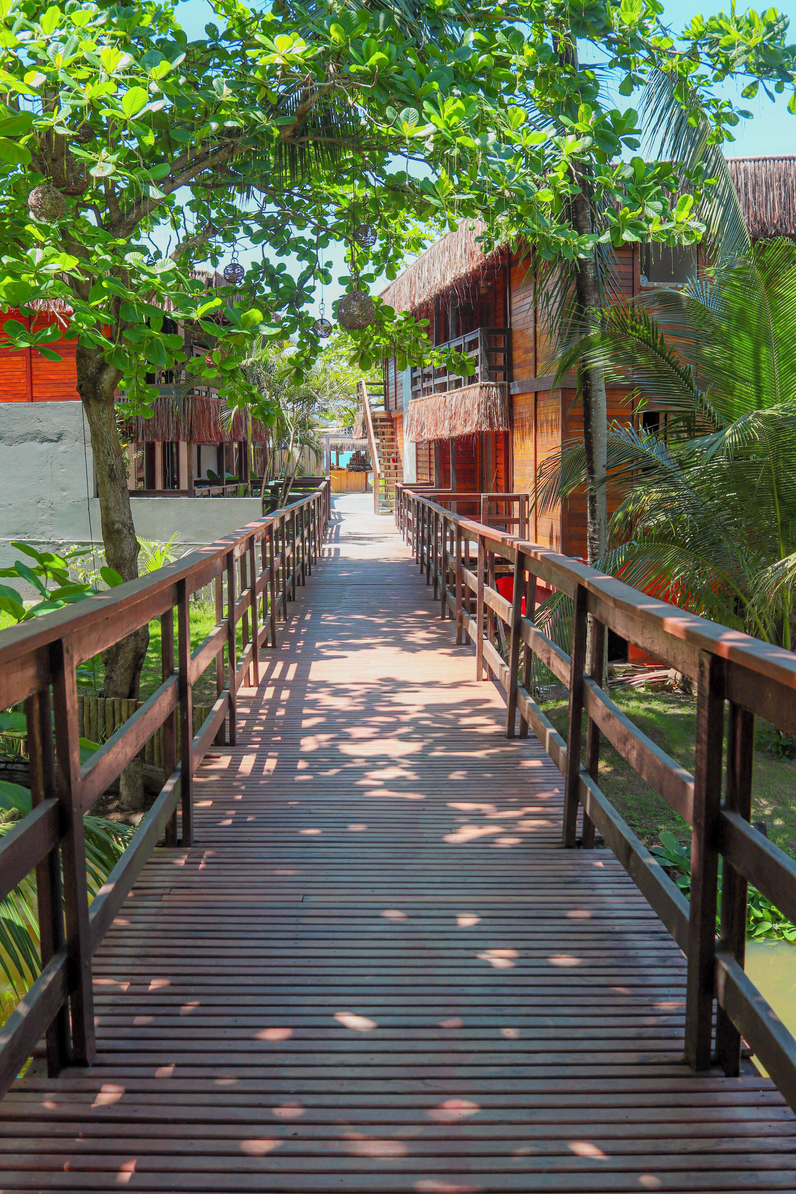 A wooden bridge leading to a building with a thatched roof, surrounded by greenery.
