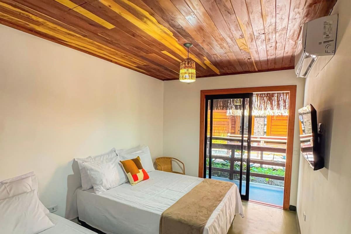 Una habitación con dos camas, techo de madera, una silla de mimbre, una lámpara en el techo y vistas a través de un ventanal.