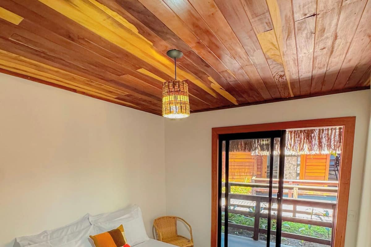 Un dormitorio con techo de madera, cama con ropa de cama blanca, lámpara de pie de fibras naturales y vistas a un balcón con mobiliario exterior.