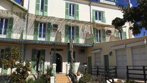 A hotel with a sign reading "HOTEL L'ILOT FLEURI". It features a two-story structure with green shutters, a balcony with a wrought iron railing, and a staircase leading to the entrance. - Hôtel L'ÎLOT FLEURI  (Le Lavandou)
