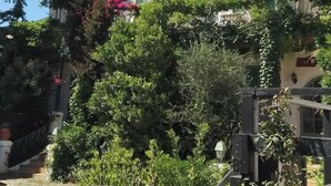 A white building with green shutters, surrounded by lush greenery and flowering plants. - Hôtel L'ÎLOT FLEURI  (Le Lavandou)