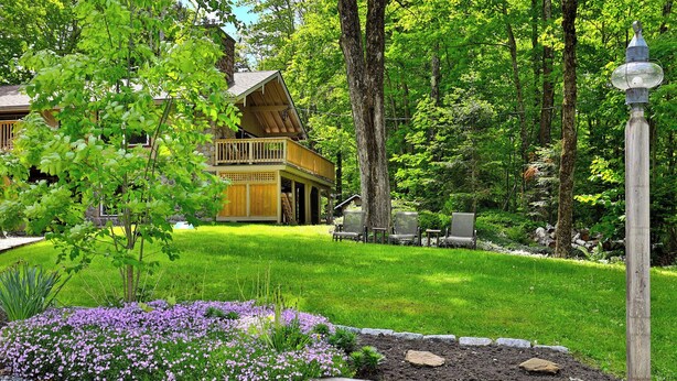 A cabin with a wooden deck, surrounded by lush greenery and a garden with purple flowers. - Fun In Fun Out Upper: 3BR with Two Decks, Sauna and Hot Tub (Killington)