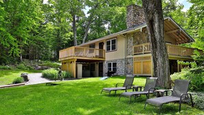 A two-story house with a stone facade, a balcony, and a grassy yard with outdoor seating. - Fun In Fun Out Upper: 3BR with Two Decks, Sauna and Hot Tub (Killington)