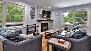 A modern living room with a fireplace, leather sofas, a coffee table, and large windows with a view of trees. - Fun In Fun Out Upper: 3BR with Two Decks, Sauna and Hot Tub (Killington)