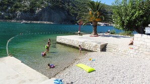 A pebbled beach with clear water, a palm tree, and a rocky hillside in the background. - Villa Riva (Dubrovnik)