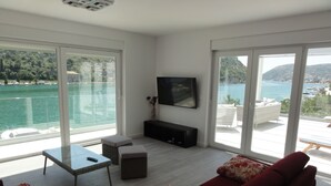 A modern living room with a large window overlooking a body of water, a flat-screen TV mounted on the wall, and a red sofa with floral cushions. - Villa Riva (Dubrovnik)