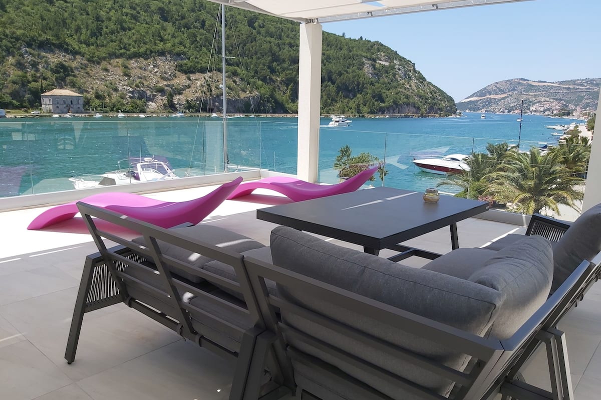 A balcony with a table and chairs, overlooking a coastal area with boats and a hilly landscape.