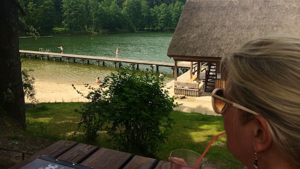 Blick auf einen See mit einem Holzsteg, einem Gebäude mit Strohdach und einer Person mit einem Getränk.