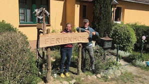 Ein Holzschild mit der Aufschrift 'FERIENHOF' vor einem gelben Gebäude mit einem Ziegeldach und grünen Fensterrahmen.