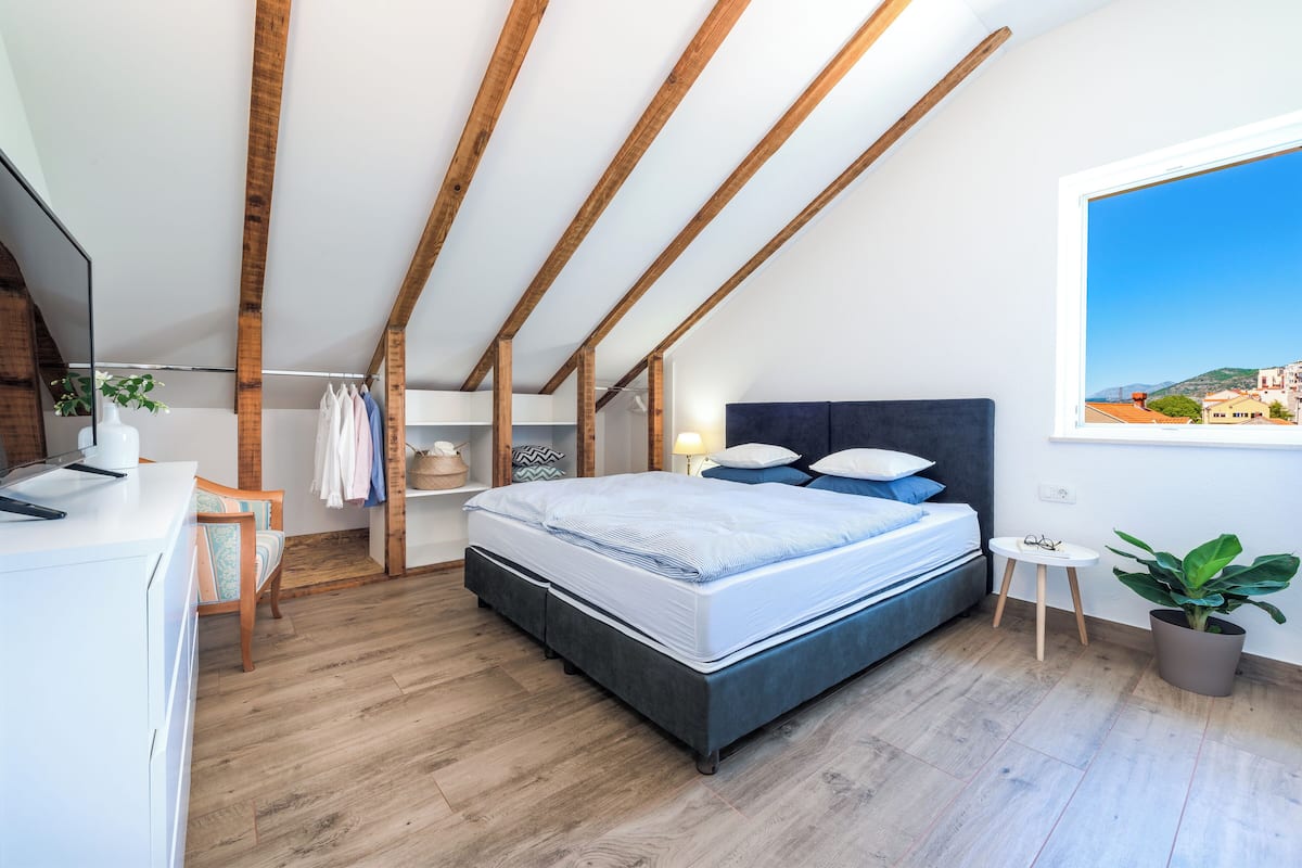 A modern bedroom with a large bed, wooden beams, a desk, and a view of a cityscape.