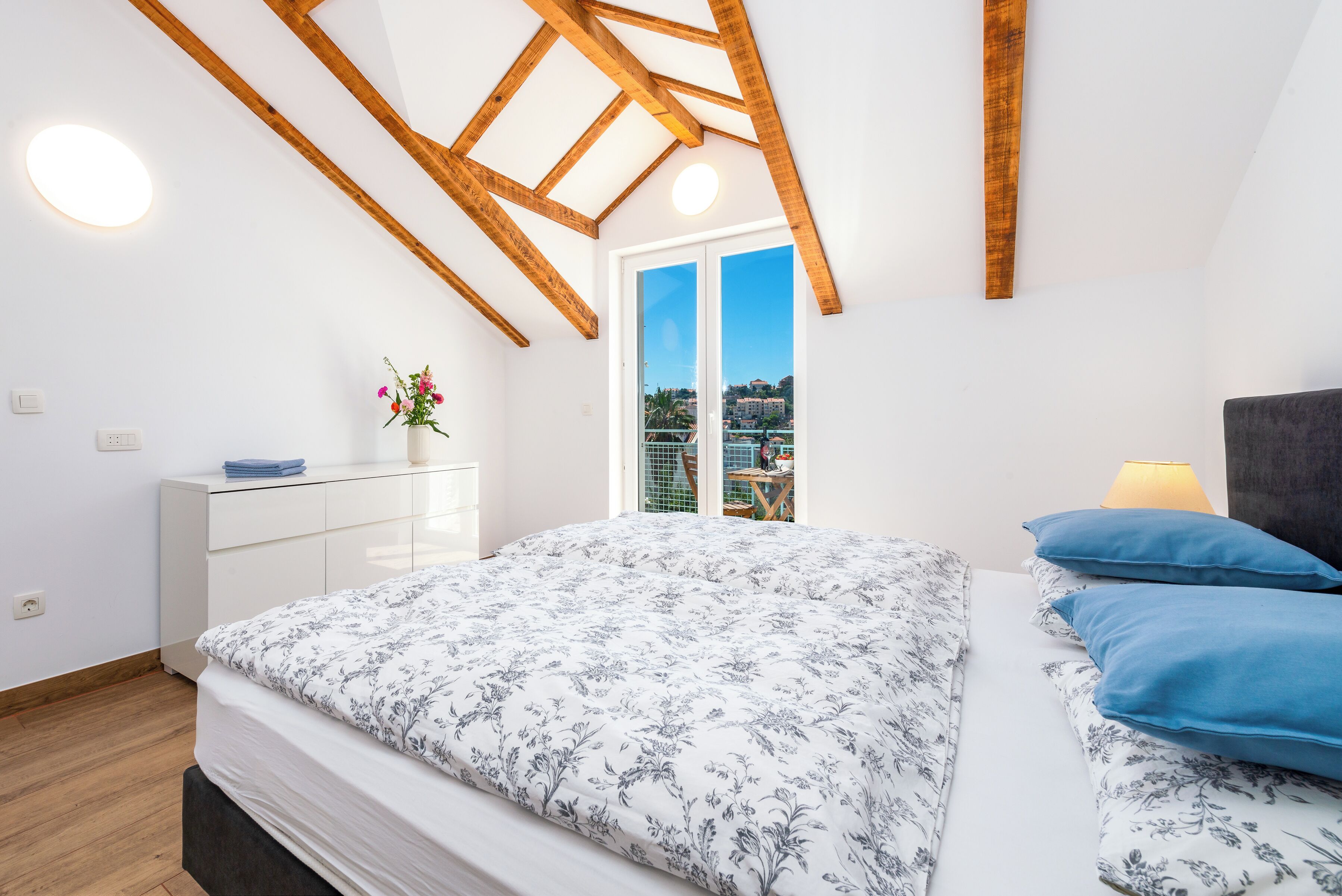 A bedroom with a bed, a dresser, a nightstand, a lamp, and a view of the outside.