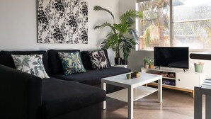 A modern living room with a black sofa, white coffee table, and a flat-screen TV on a white stand. There are several potted plants and a large window with a view of trees. - Headlands 9 - Forster beachfront (Forster)