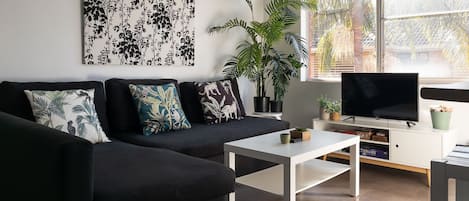 A modern living room with a black sofa, white coffee table, and a flat-screen TV on a white stand. There are several potted plants and a large window with a view of trees.