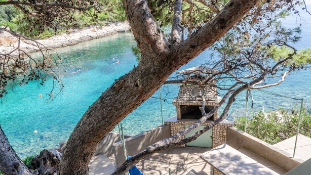 A stone-built outdoor seating area with a view of a clear blue lake, surrounded by trees and a stone wall. - Seafront Apartments Olivera (Blato)