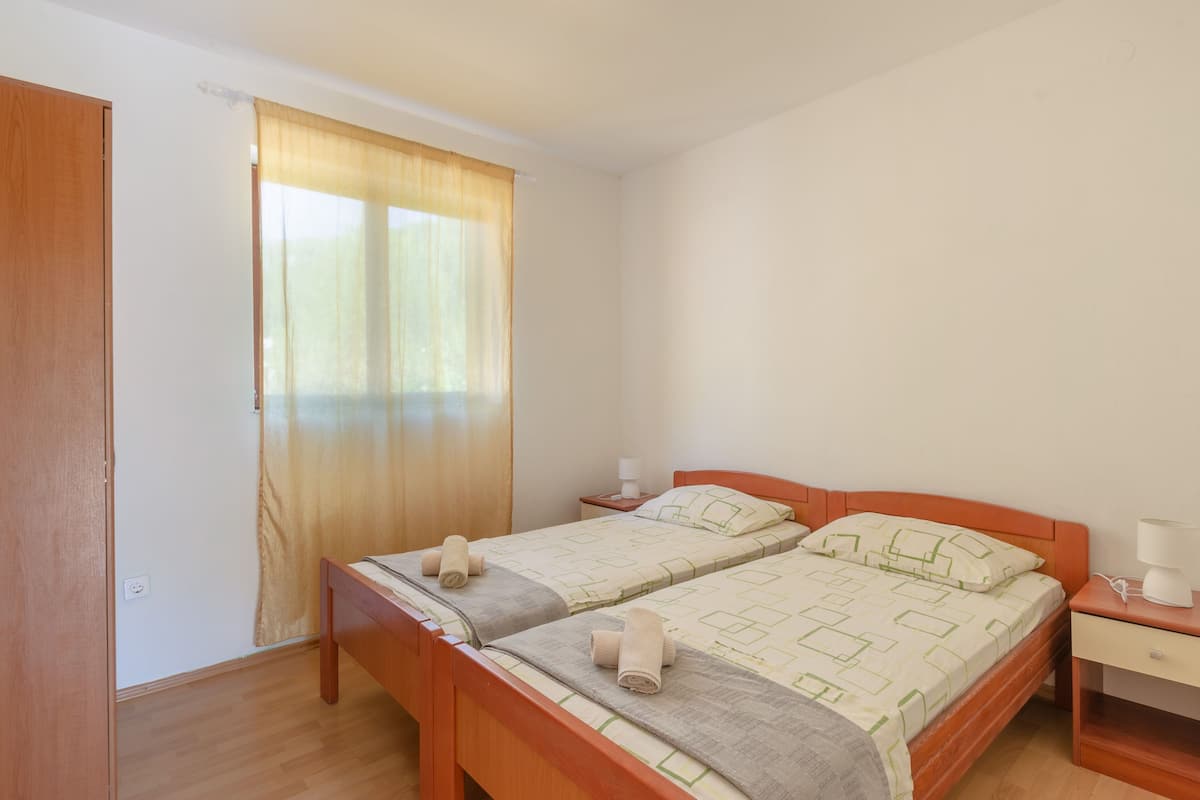 A room with two wooden beds, a wooden wardrobe, a window with curtains, and a bedside table with a lamp.