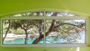 A room with a large window overlooking a turquoise body of water, surrounded by trees. - Seafront Apartments Olivera (Blato)